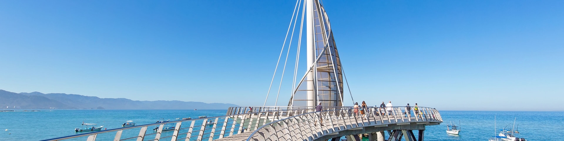 Un pont moderne au design original, qui enjambe une mer calme où l’on aperçoit des bateaux.