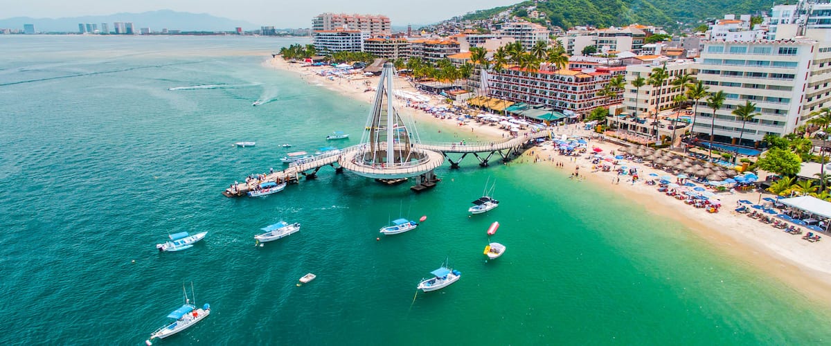 A coastal city with a prominent bridge connecting two land masses, a beach with umbrellas, and several boats in the water.