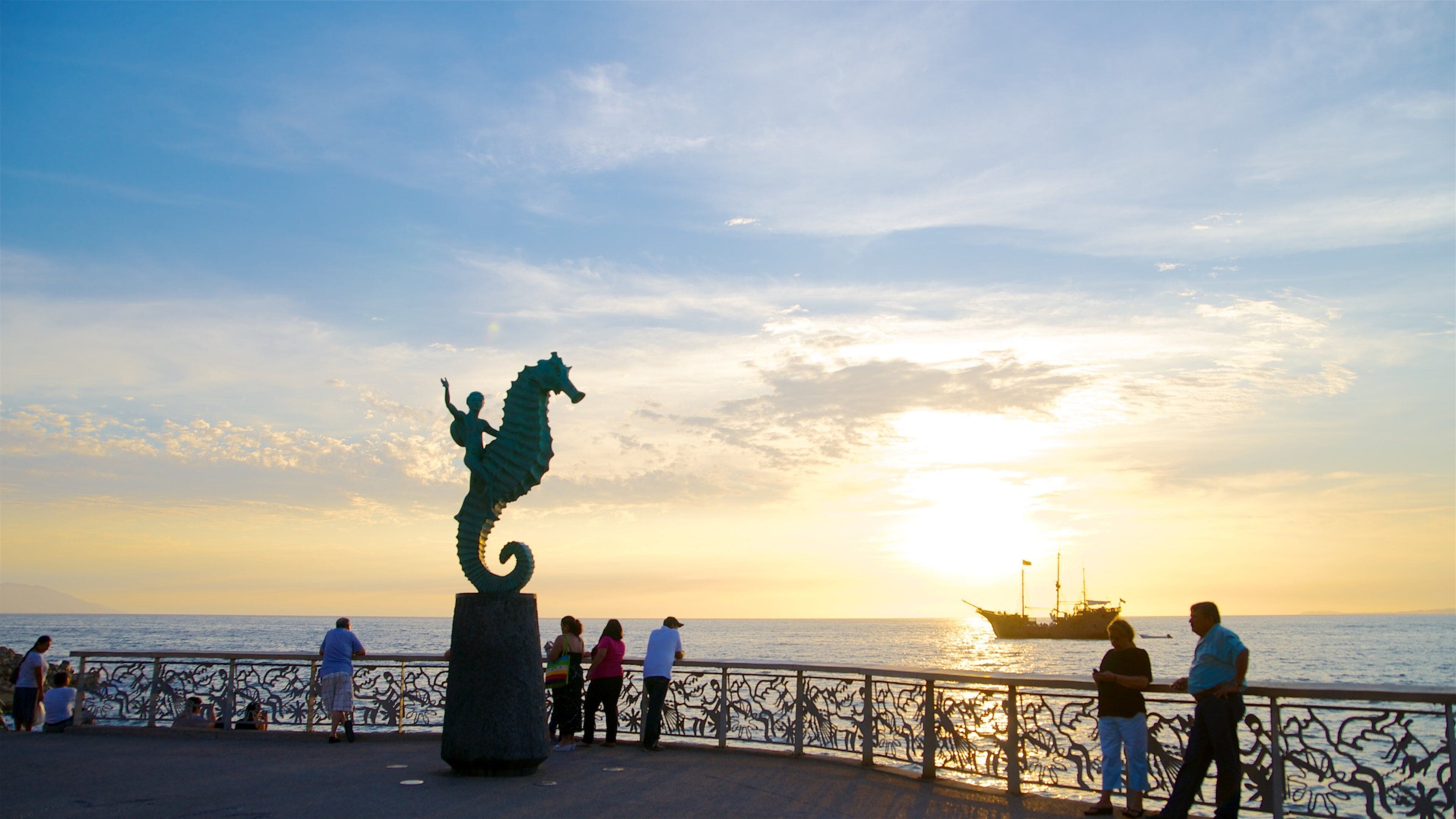 Una escultura de caballito de mar en un muelle, con gente observando la puesta de sol y un barco a lo lejos.