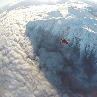 A person skydiving over a mountainous landscape with snow-covered peaks and clouds below.