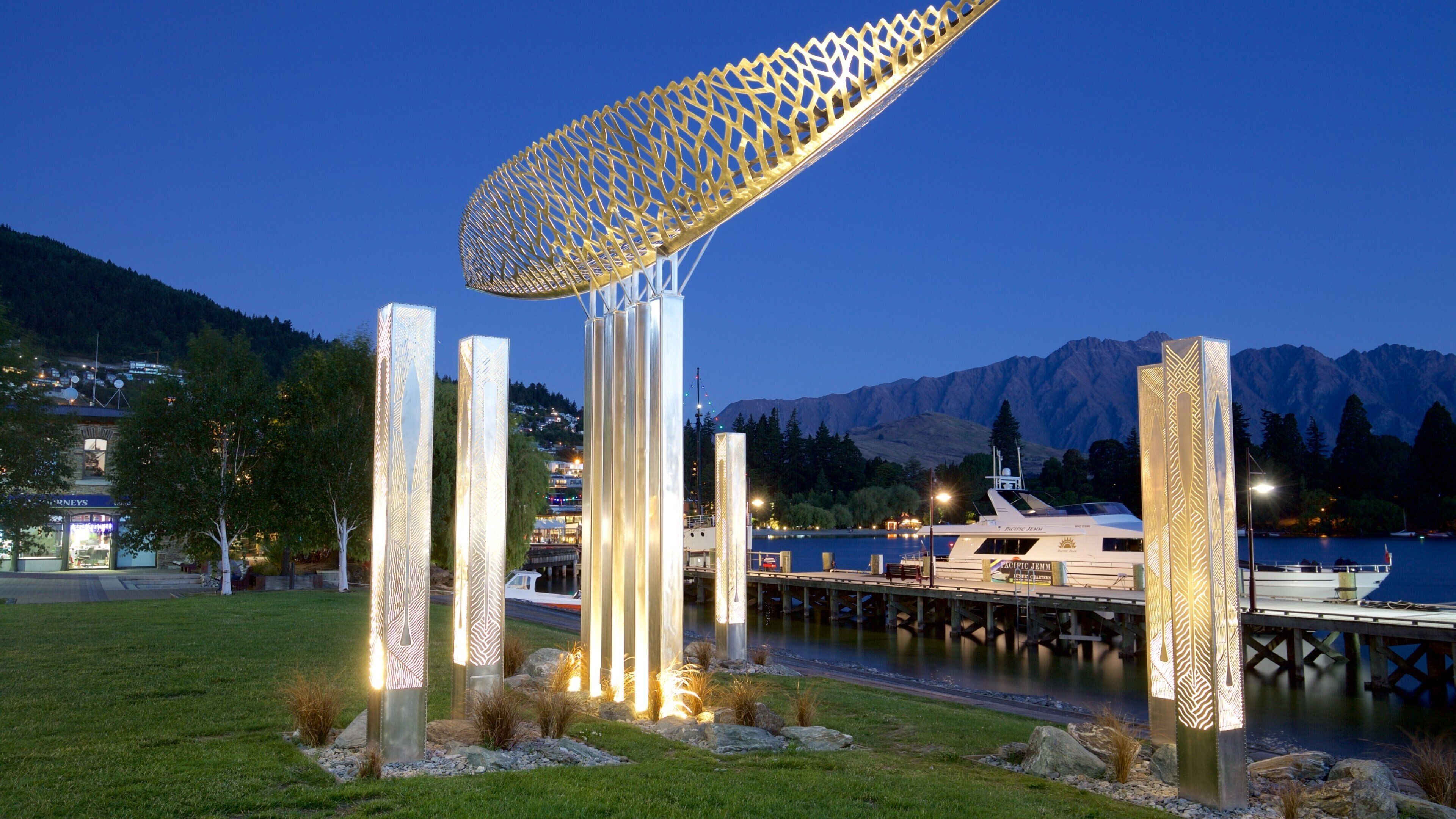Escultura moderna com uma cobertura geométrica, cercada por pilares iluminados, com um lago e montanhas ao fundo ao entardecer.