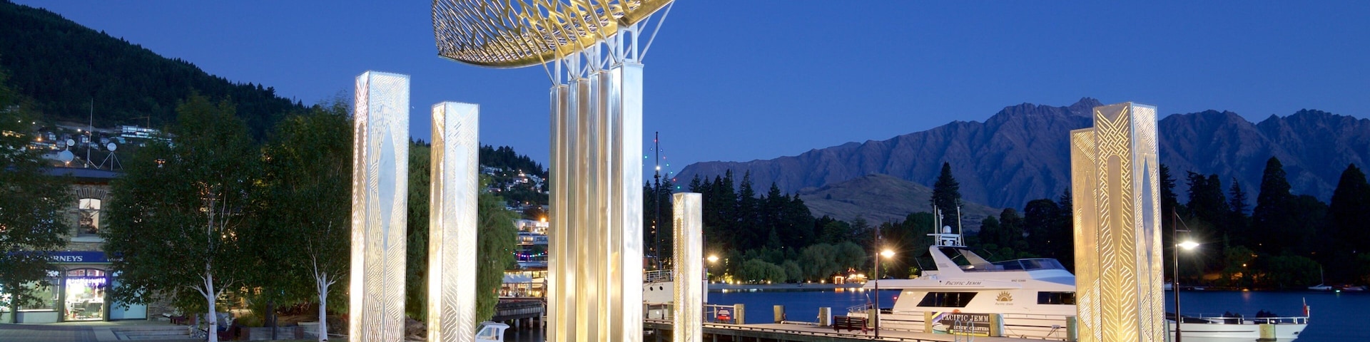 Escultura moderna com uma cobertura geométrica, cercada por pilares iluminados, com um lago e montanhas ao fundo ao entardecer.