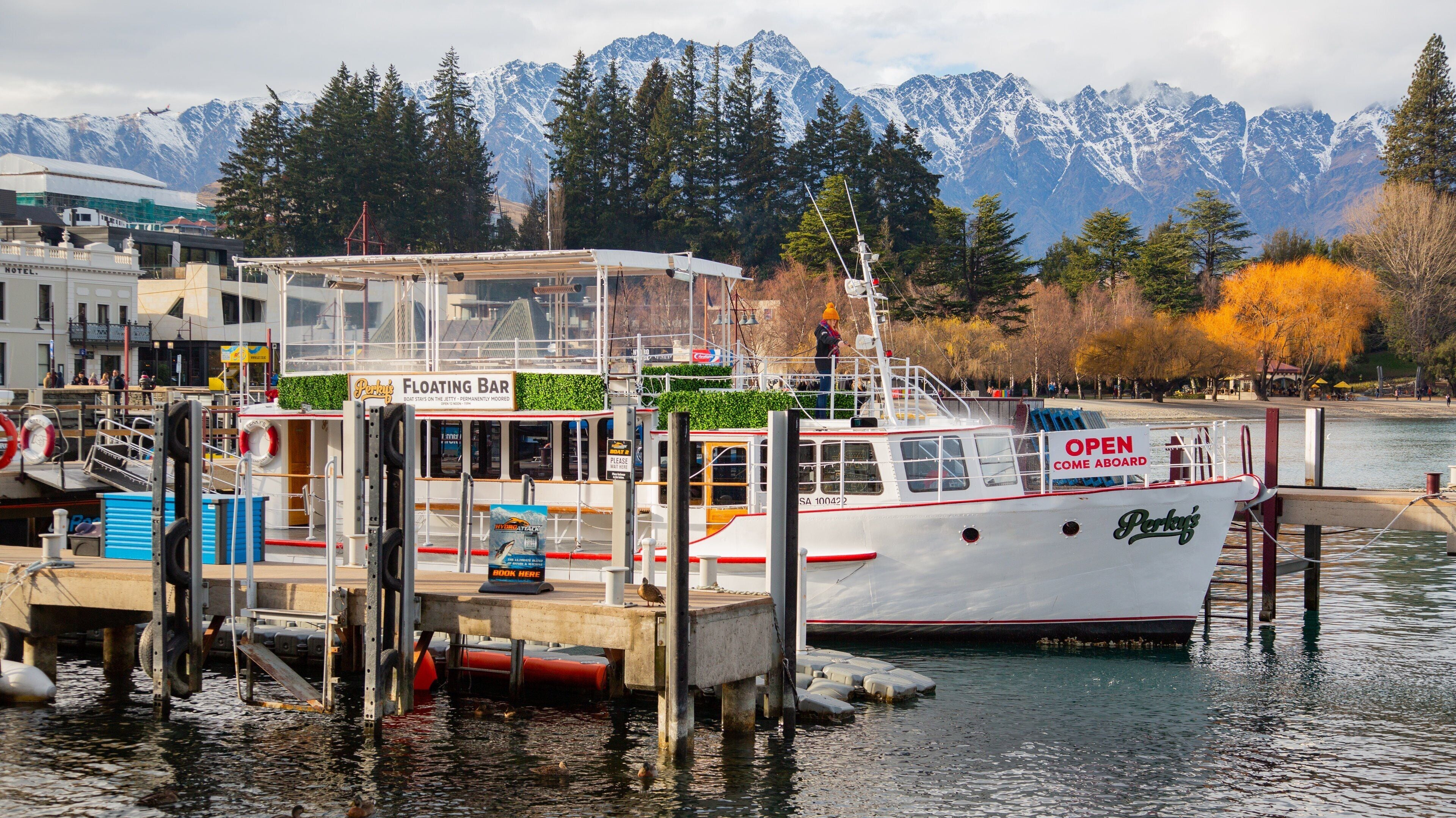 A white boat docked at a pier with a floating bar named "Perky" and a sign that says "OPEN COME ABOARD".