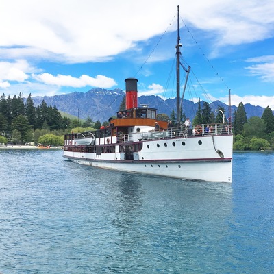 A white steamboat with a red funnel and a cabin, sailing on a calm river with mountains in the background.