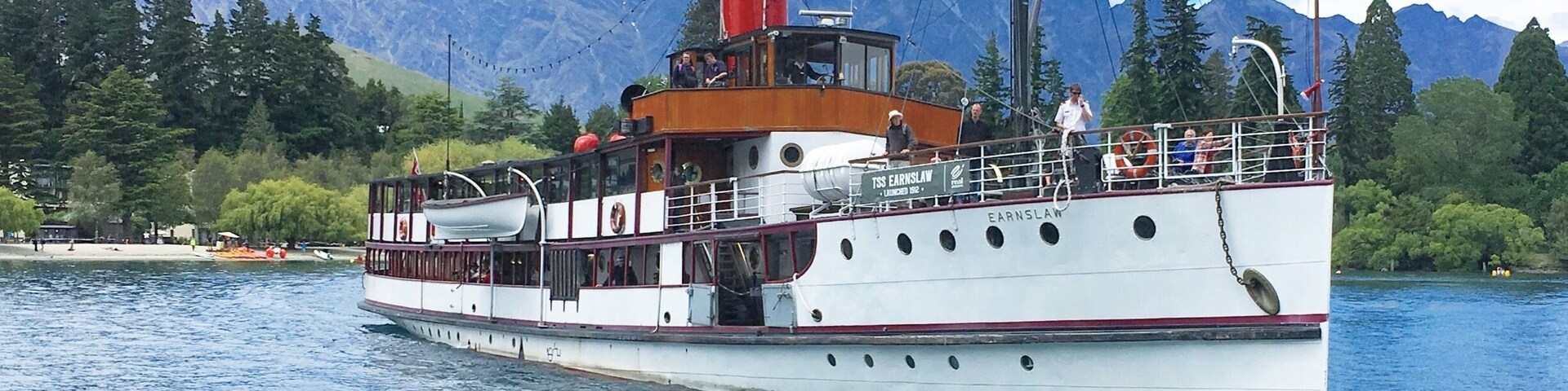 Um barco a vapor branco, com chaminé vermelha e cabine, navega em um rio calmo, com montanhas ao fundo.