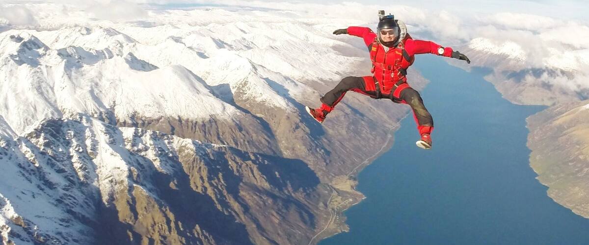 A person skydiving over a mountainous landscape with snow-covered peaks and a body of water below.