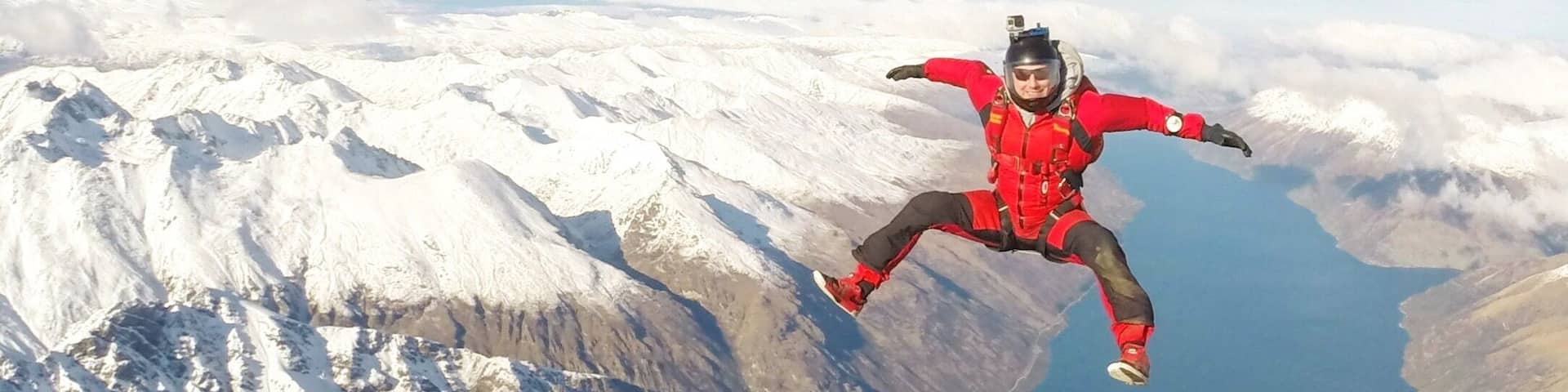 A person skydiving over a mountainous landscape with snow-covered peaks and a body of water below.