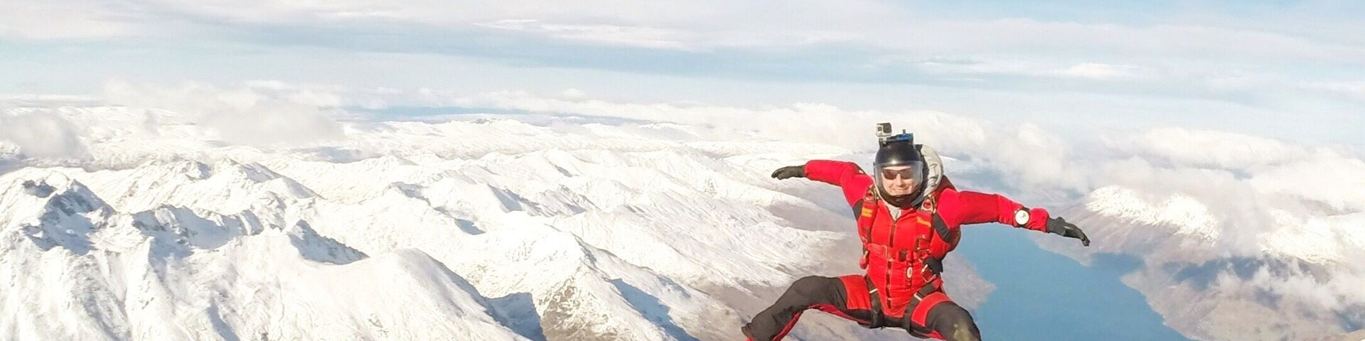 Uma pessoa praticando paraquedismo sobre uma paisagem montanhosa com picos cobertos de neve e um corpo d'água abaixo.