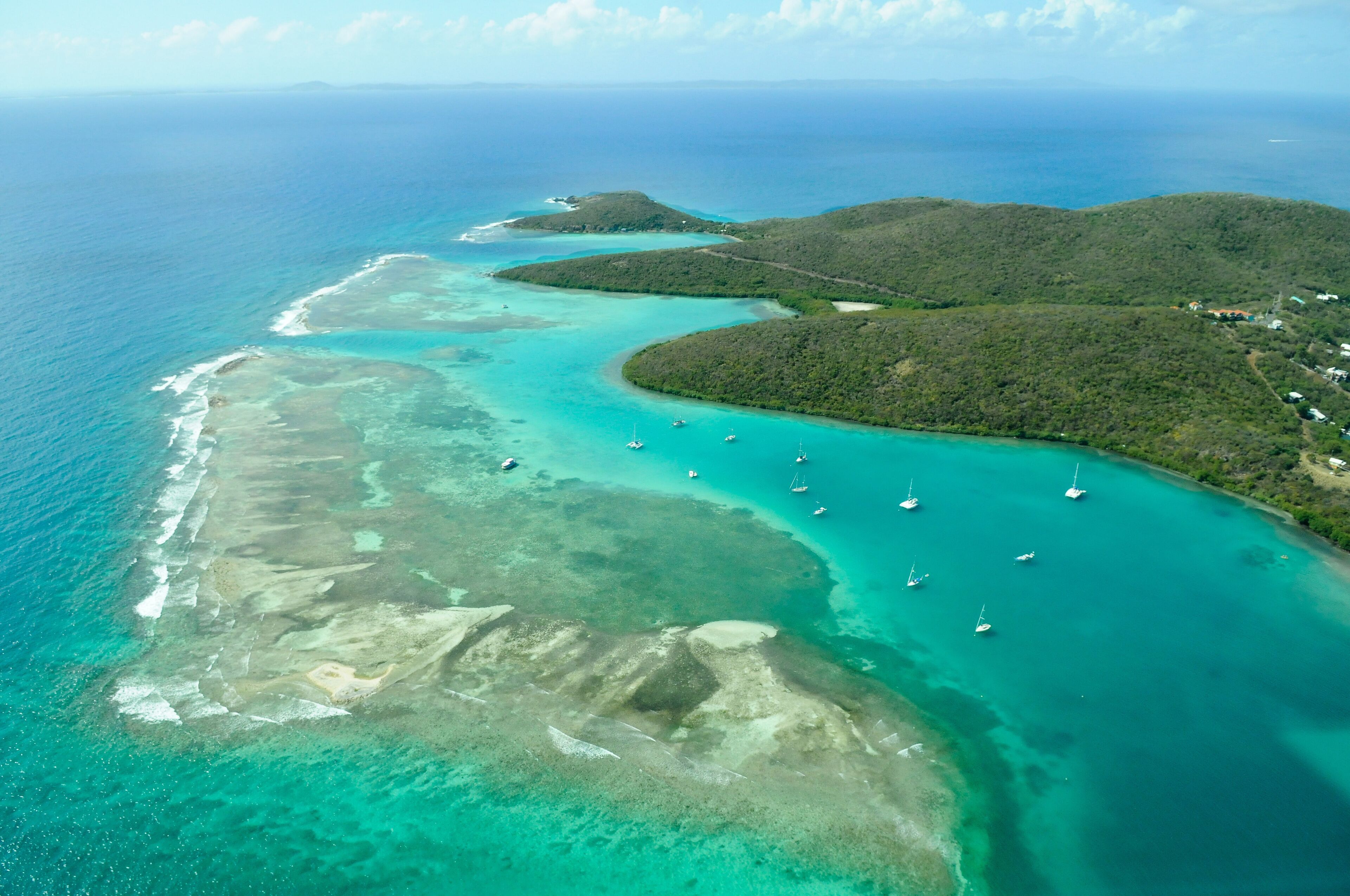 An aerial view of a turquoise coastal area with clear waters, small islands, and boats anchored near the shore.