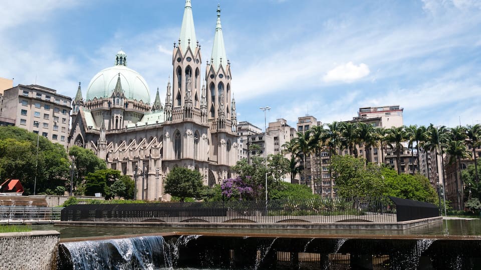A cathedral with twin spires and a large dome, a water feature, and surrounding palm trees.