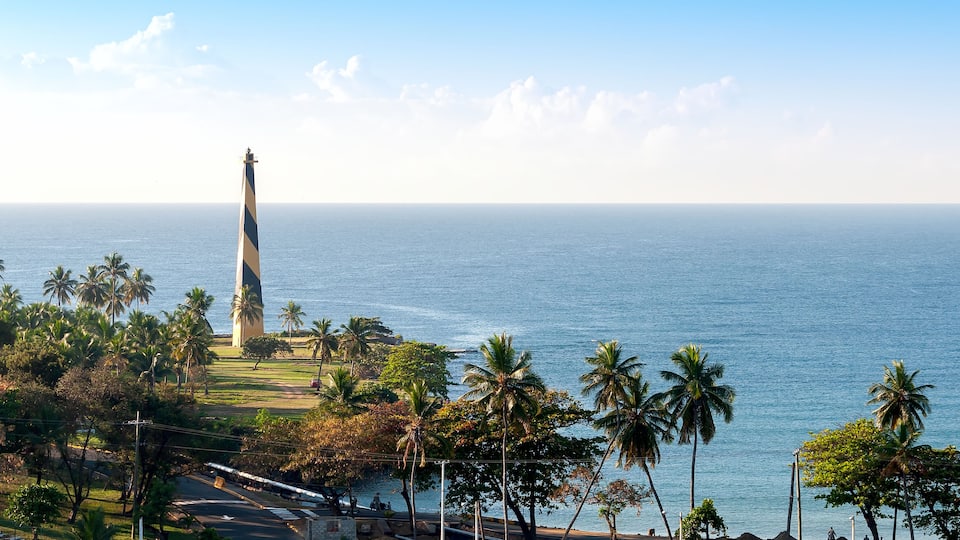 A coastal landscape with a lighthouse, palm trees, and a clear view of the ocean.