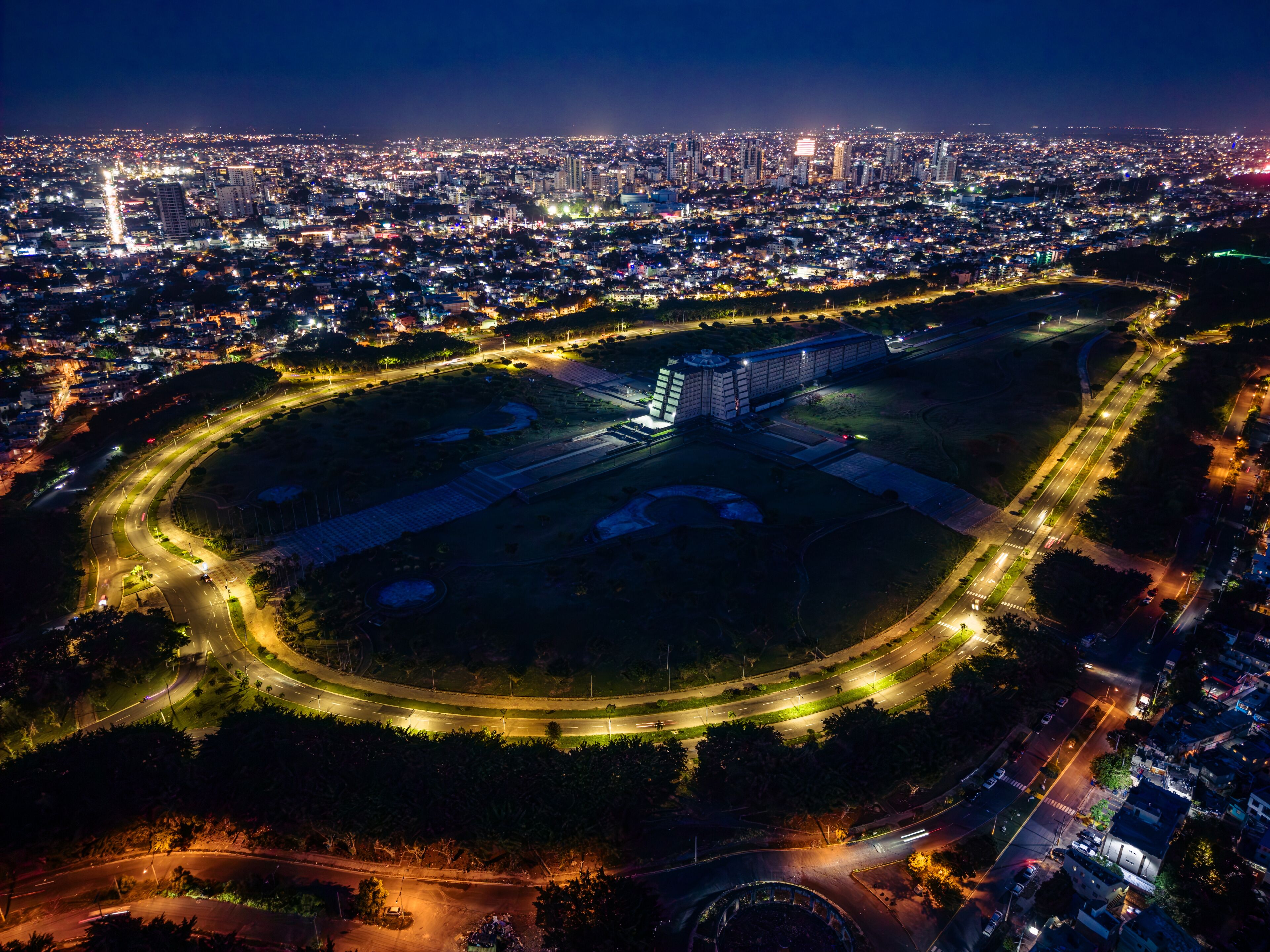 A cityscape at night with a large, illuminated sports stadium surrounded by a well-lit road network.