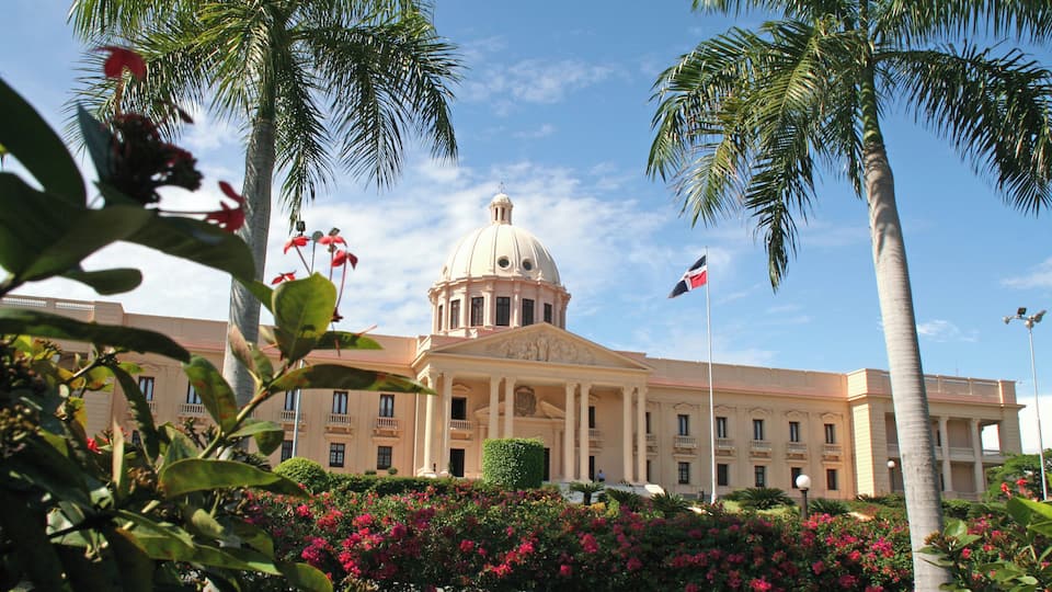 A large building with a dome and columns, surrounded by palm trees and pink flowers.