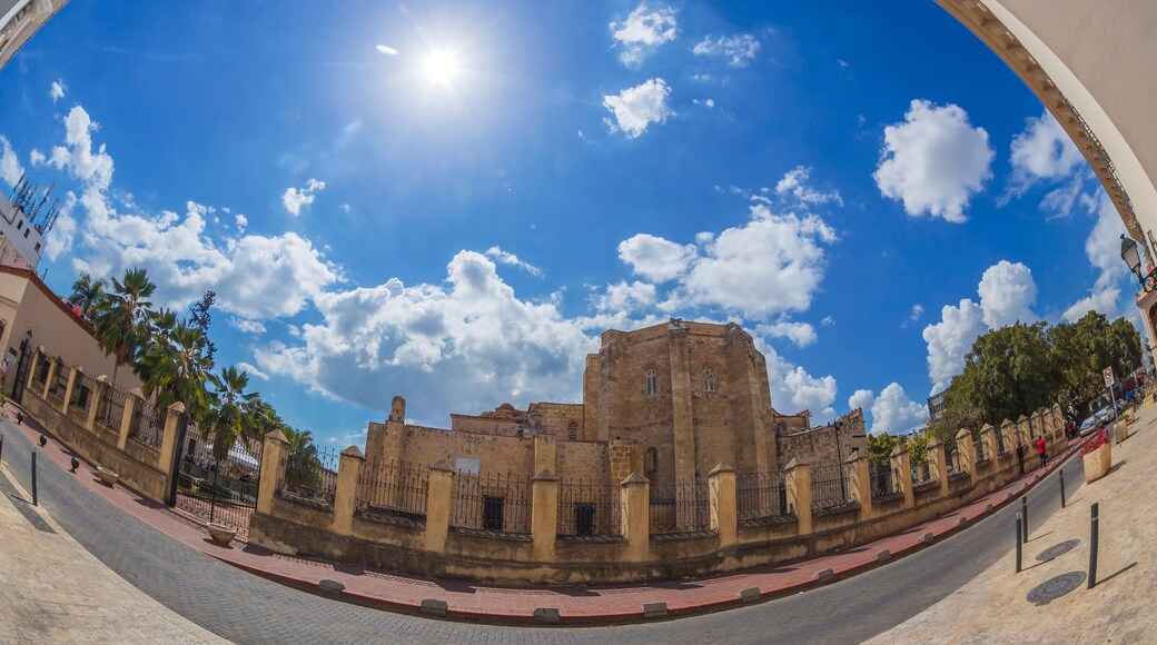 A historic building with a large central tower, surrounded by a paved area and palm trees.
