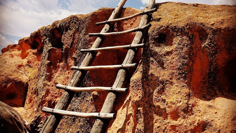 Eine Holzleiter lehnt an einem felsigen Felsen unter einem teils bewölkten Himmel.