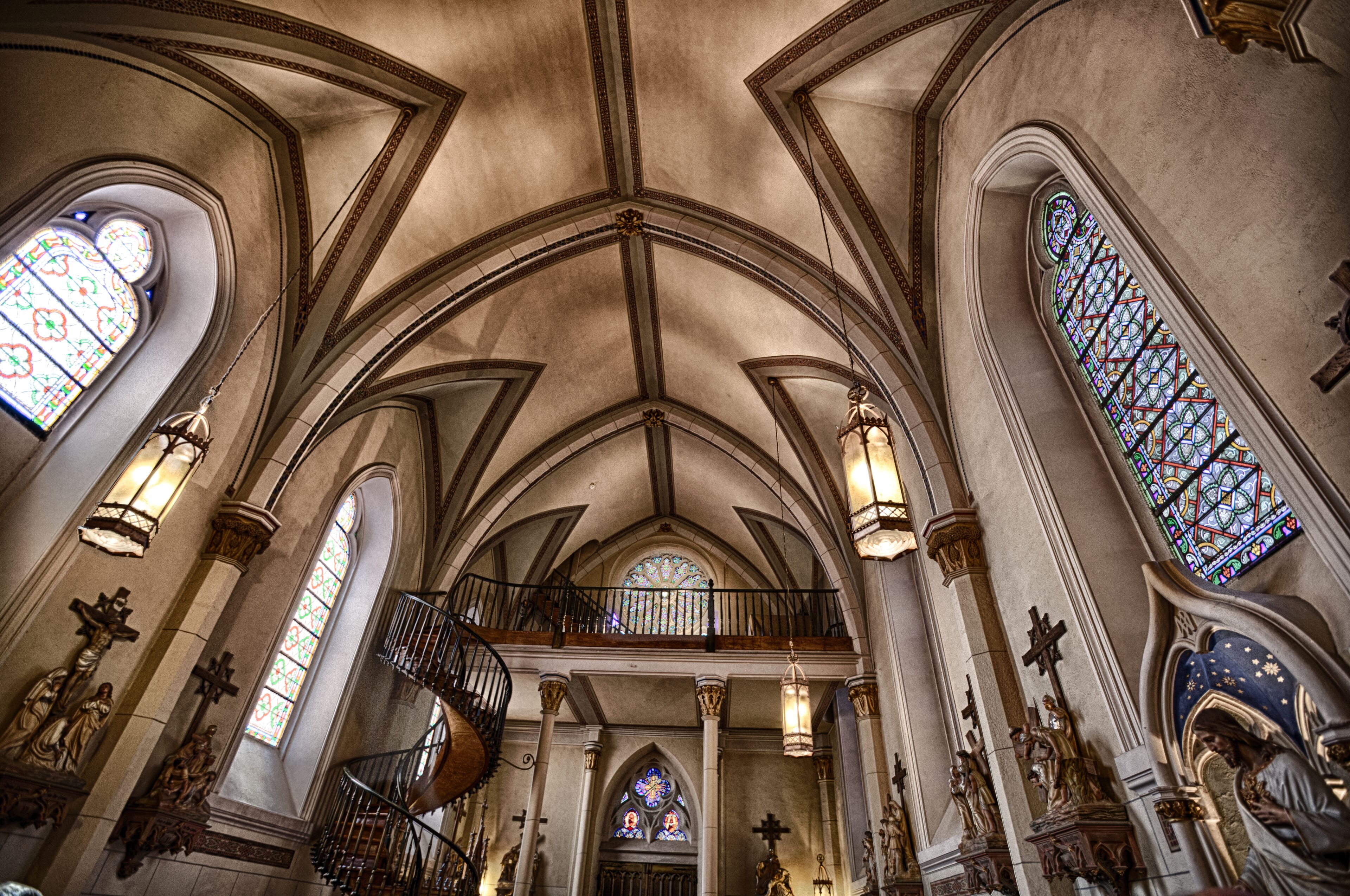 A church interior with arched ceilings, stained glass windows, and a spiral staircase.