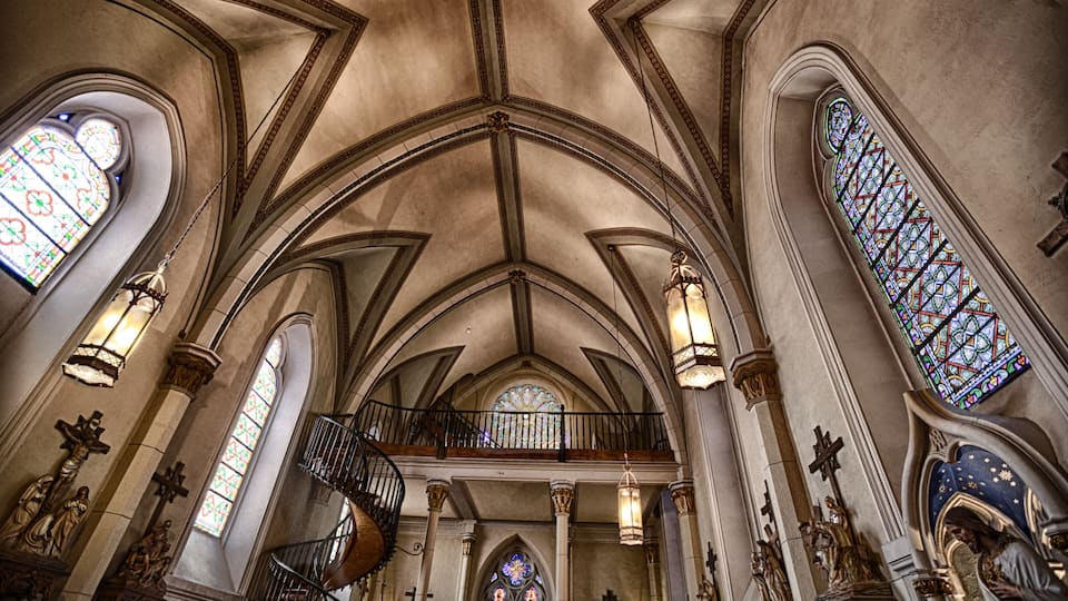 A church interior with arched ceilings, stained glass windows, and a spiral staircase.