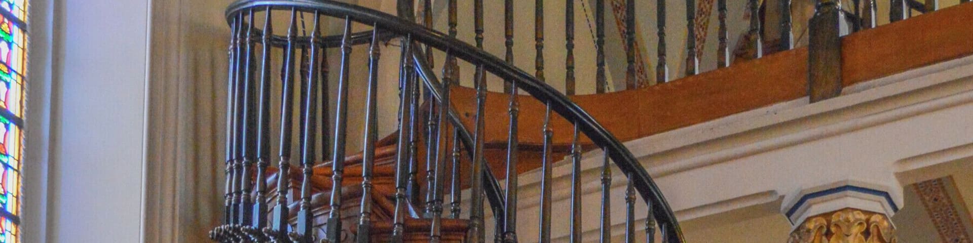 A grand spiral staircase with ornate railings and wooden steps inside a building with stained glass windows and religious statues.