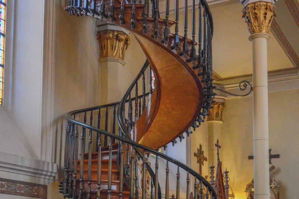 A grand spiral staircase with ornate railings and wooden steps inside a building with stained glass windows and religious statues.