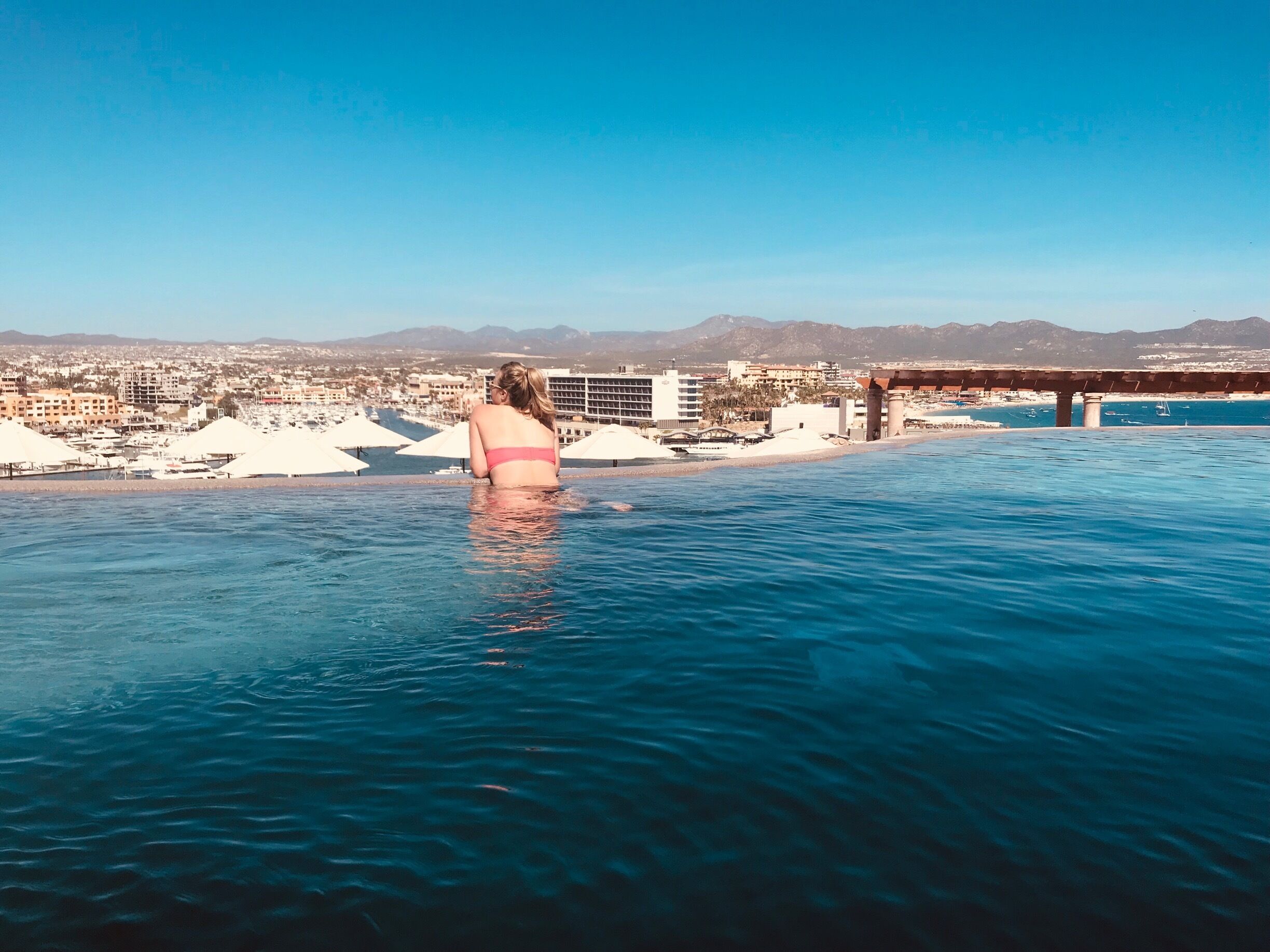 A woman in a pink swimsuit at an infinity pool with a cityscape and mountains in the background.