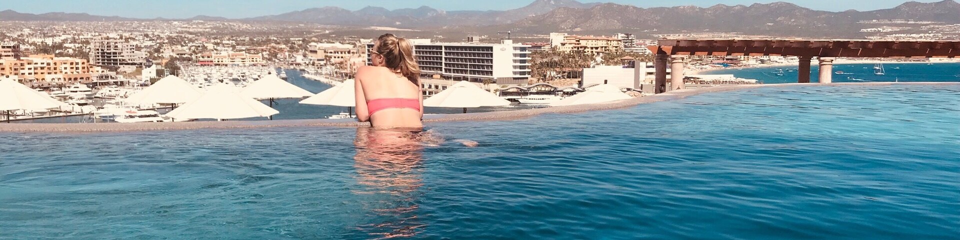 A woman in a pink swimsuit at an infinity pool with a cityscape and mountains in the background.