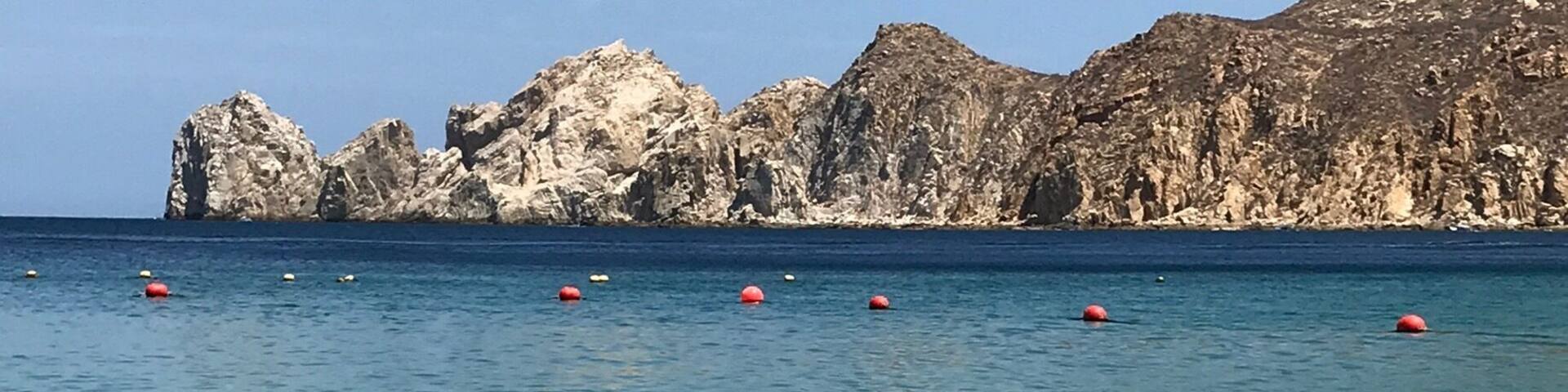 A clear coastal beach with shallow water, rocky cliffs, and several buoys in the distance.