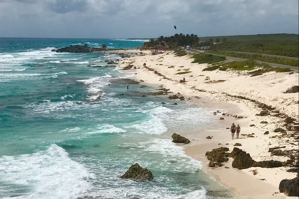 Una playa con olas rompiendo en la orilla, algunas personas caminando y un camino que lleva a un pueblo lejano.