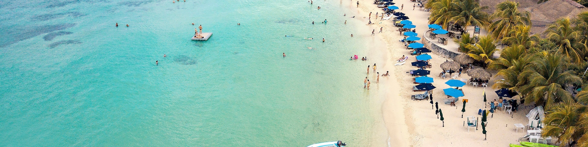 Uma praia tropical com águas turquesa cristalinas, uma fileira de guarda-sóis e um barco ancorado perto da costa.