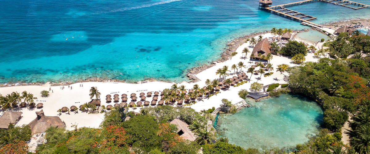 A tropical resort with a white sandy beach, palm trees, clear turquoise waters, and a pier extending into the sea.
