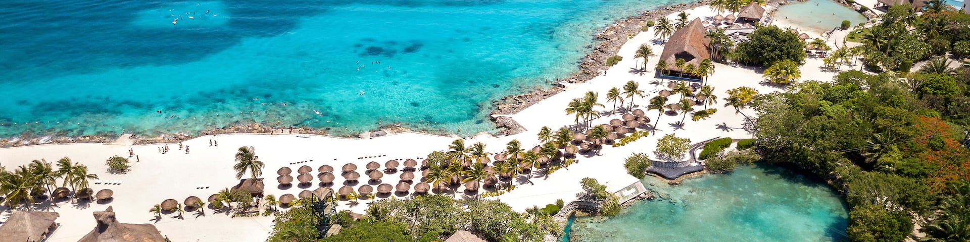 A tropical resort with a white sandy beach, palm trees, clear turquoise waters, and a pier extending into the sea.