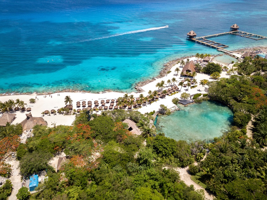Um resort tropical com uma praia de areia branca, palmeiras, águas turquesa cristalinas e um píer que se estende para o mar.