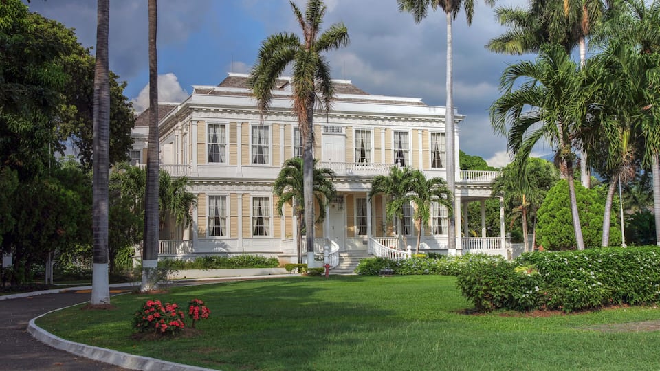 A large, historic building with a white facade and columns, surrounded by lush greenery and palm trees.