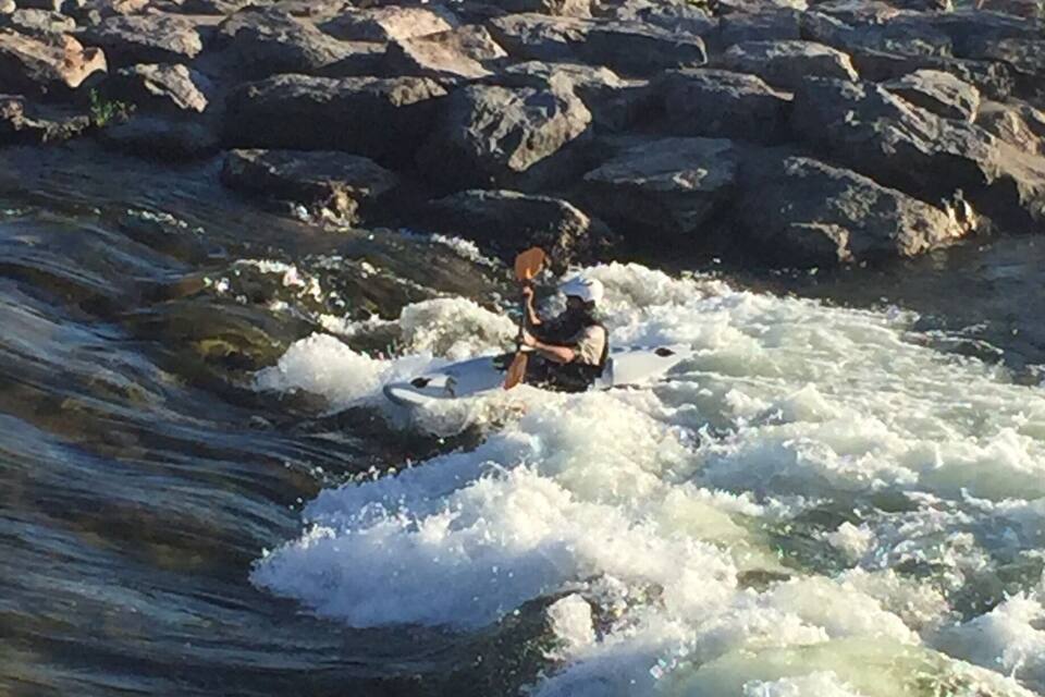 Un remer de kayak navegant per un riu amb vores rocoses i altres kayakistes al fons.