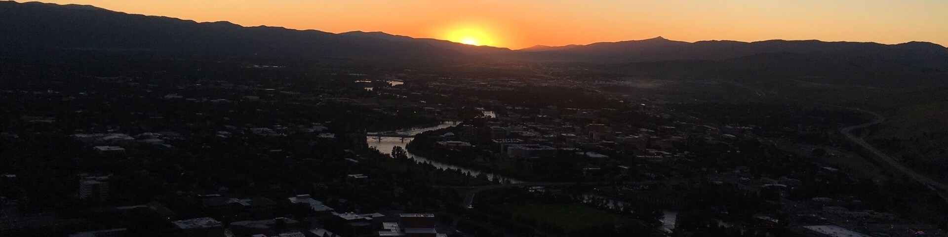 A sunset over a valley with a river, buildings, and mountains in the background.