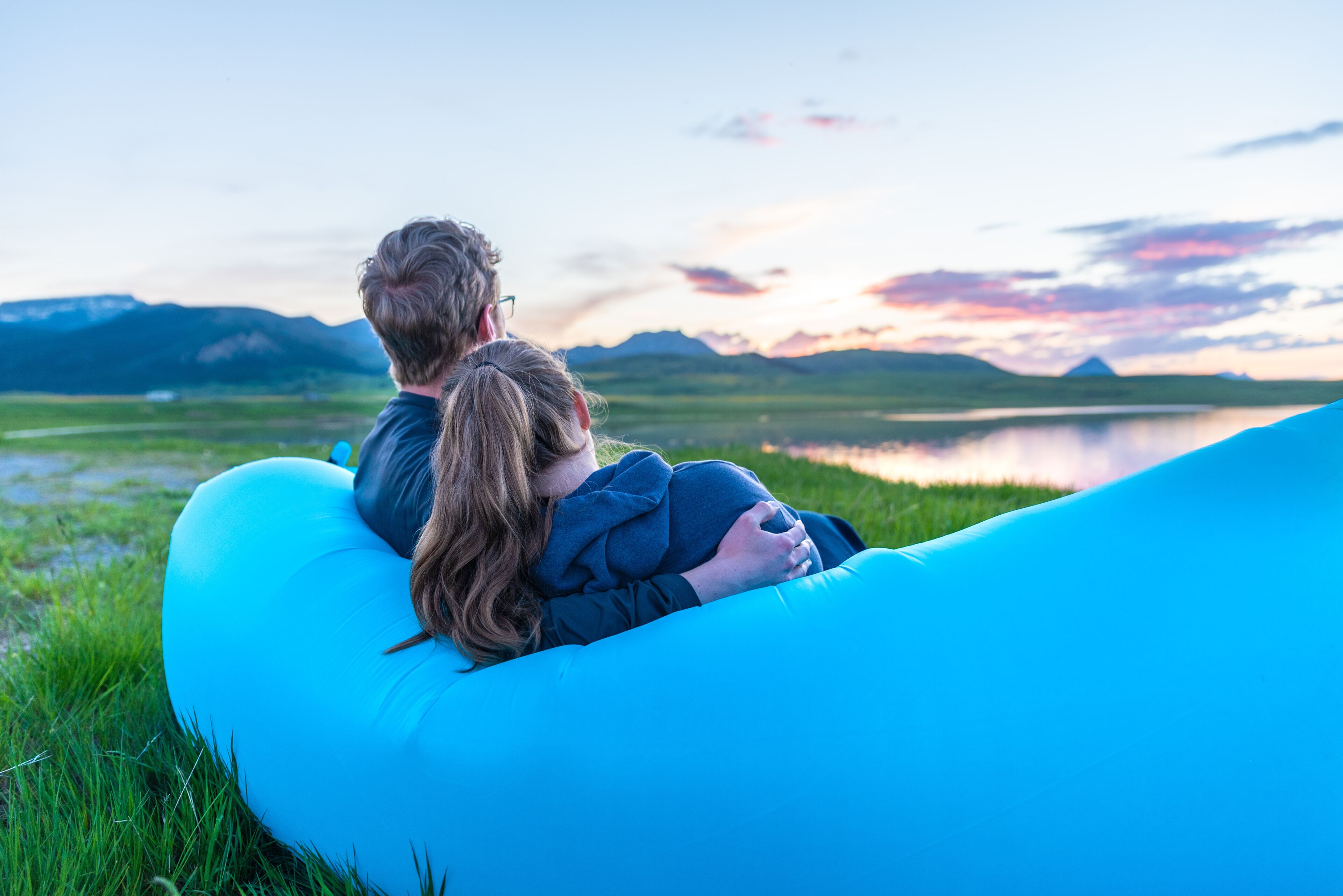 Una pareja sentada sobre un cojín inflable azul junto a un lago, con montañas al fondo.