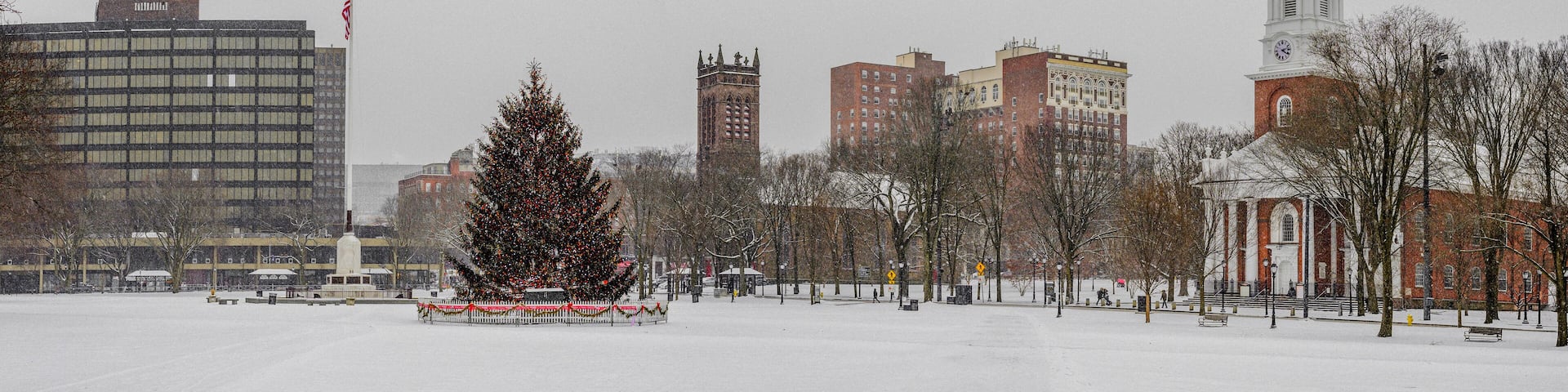 A snow-covered park with a decorated Christmas tree, a church with a steeple, and several buildings in the background.