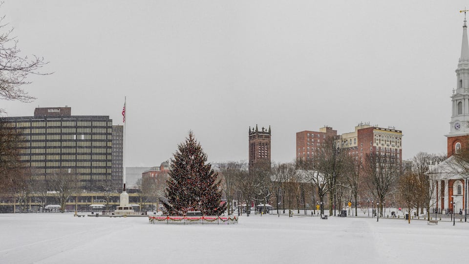 A snow-covered park with a decorated Christmas tree, a church with a steeple, and several buildings in the background.