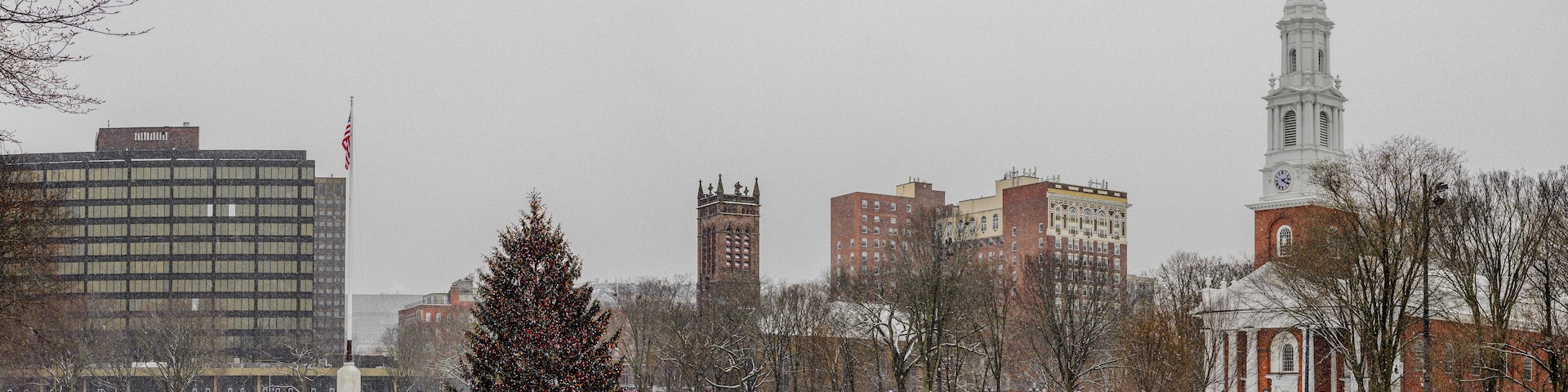 A snow-covered park with a decorated Christmas tree, a church with a steeple, and several buildings in the background.