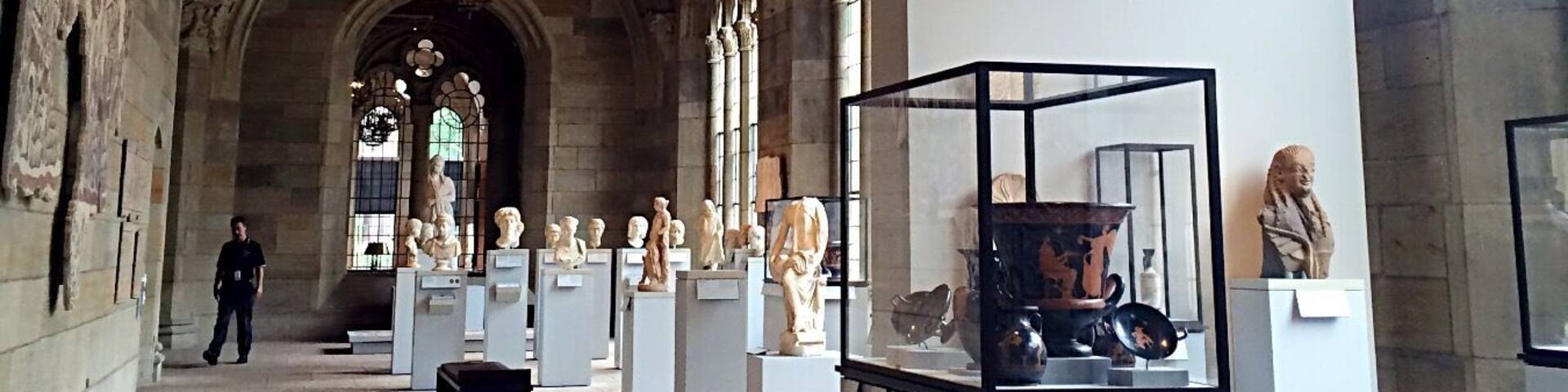 A museum interior with arched ceilings, hanging chandeliers, and classical sculptures on display.