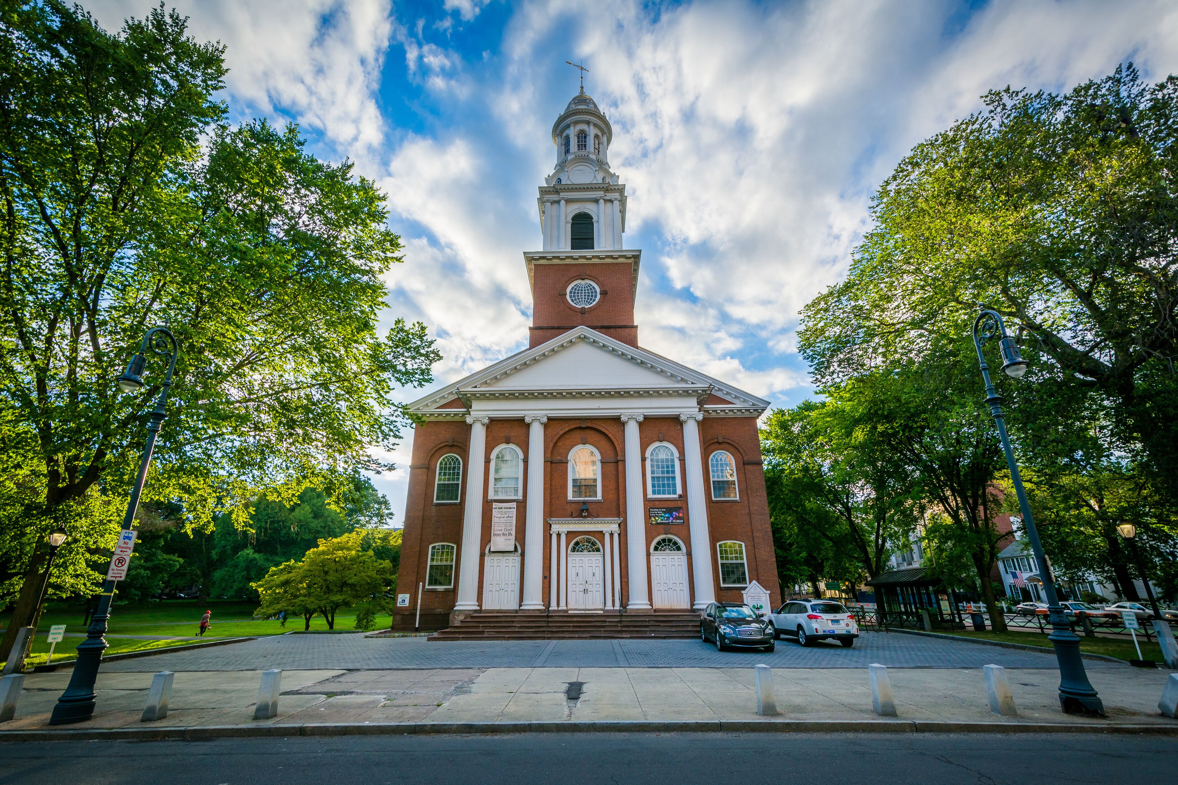 A red brick church with a white steeple and a clock tower, surrounded by trees and a paved street.