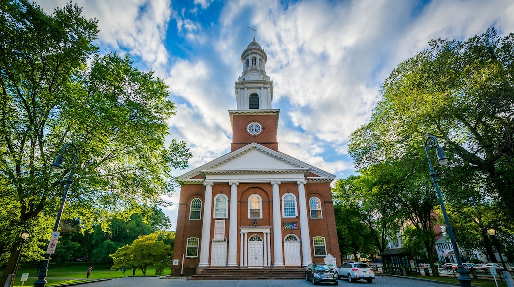 A red brick church with a white steeple and a clock tower, surrounded by trees and a paved street.
