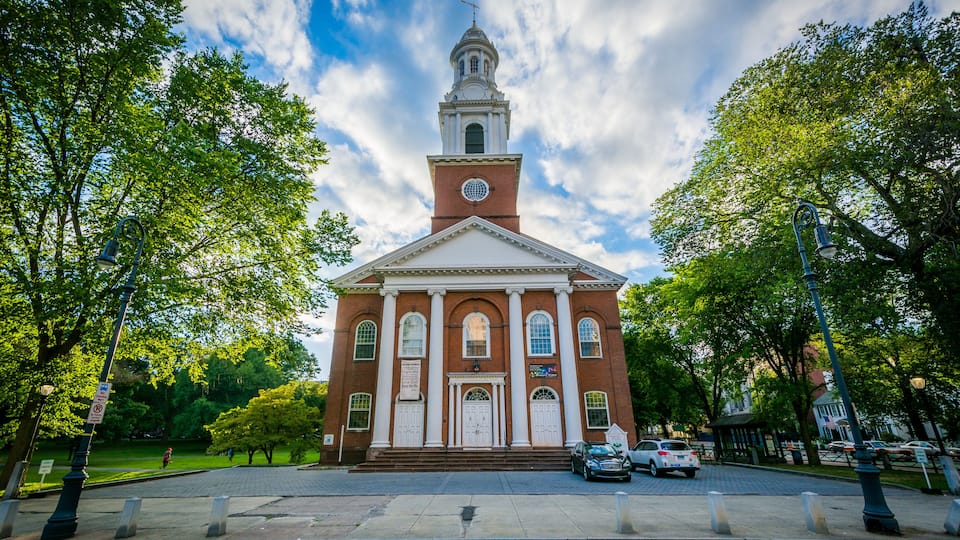 A red brick church with a white steeple and a clock tower, surrounded by trees and a paved street.