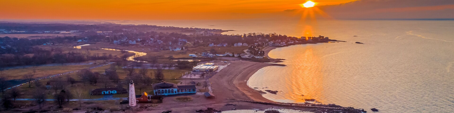 Atardecer costero con un faro, playa y un pueblo al fondo.