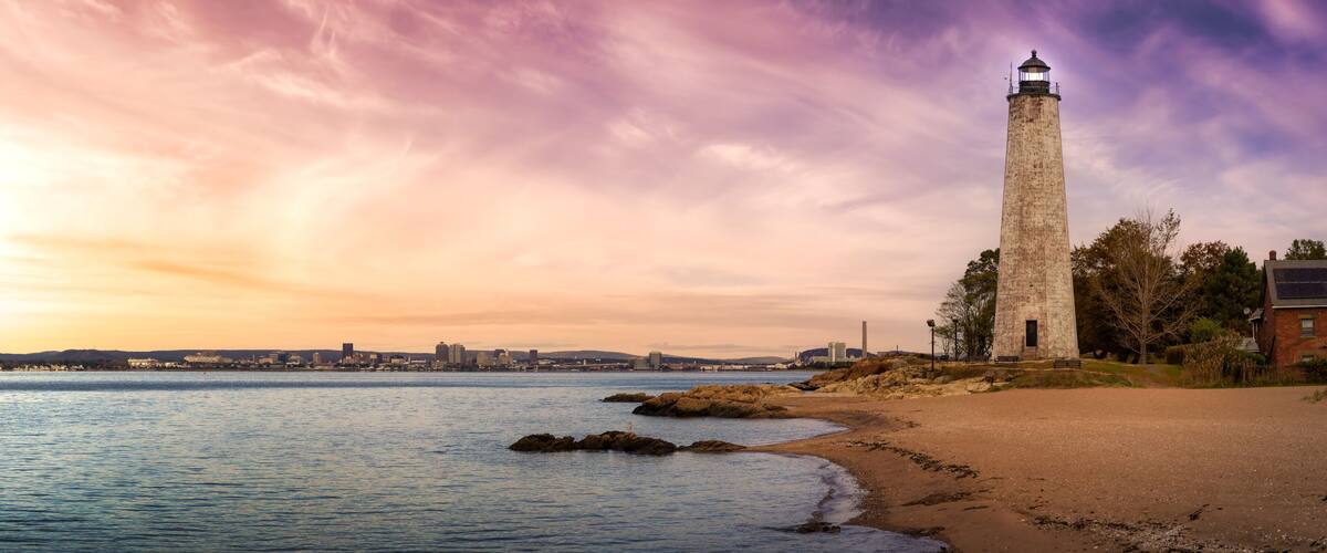 A coastal landscape with a lighthouse, beach, rocky outcrops, and a distant city skyline.