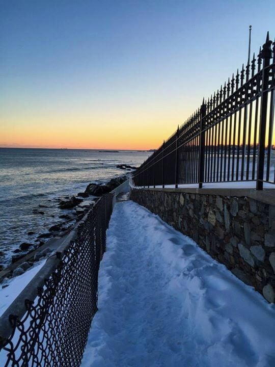 A snow-covered walkway beside a stone wall and metal fence overlooking the ocean at sunset.