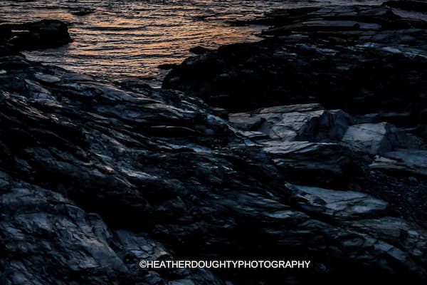 A sunset over a rocky shoreline with calm water reflecting the sky.