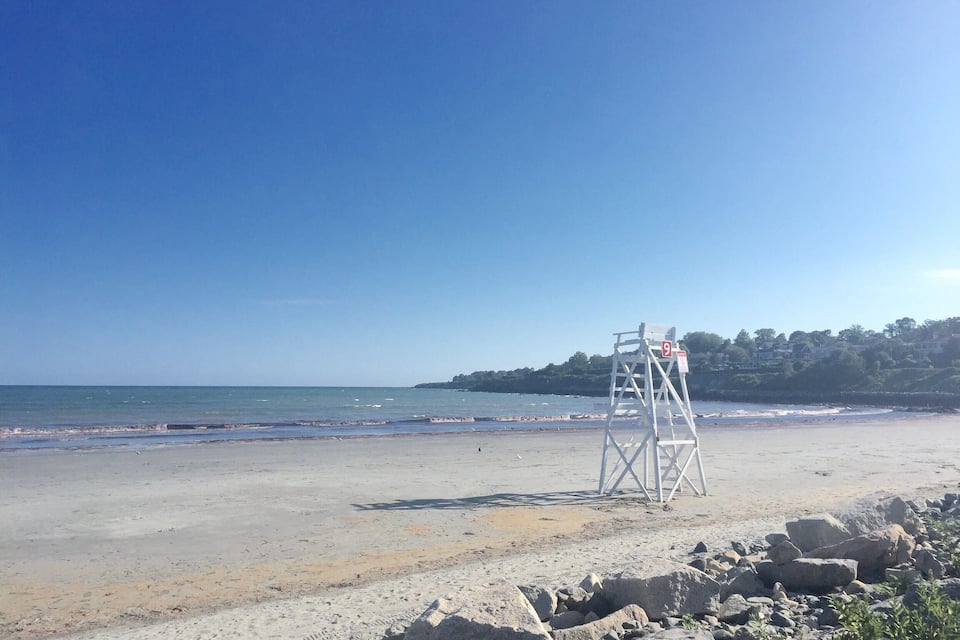 Ein Strand mit einem Rettungsturm, felsigem Ufer und einer entfernten Anhöhe mit Häusern.