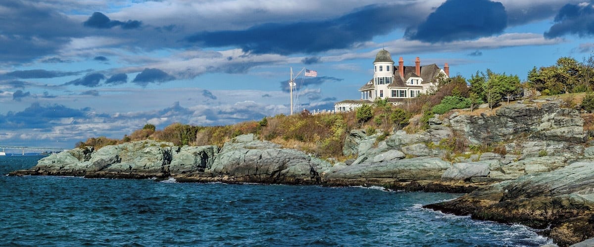 A coastal house on a rocky cliff with a view of the ocean and a wind turbine in the distance.