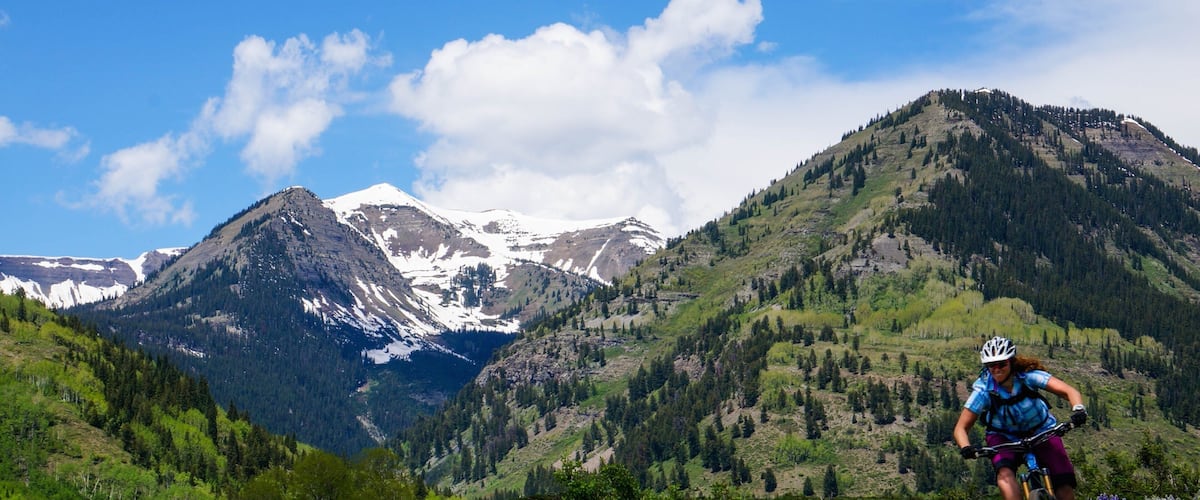 A mountain biker on a trail with snow-capped peaks in the background.