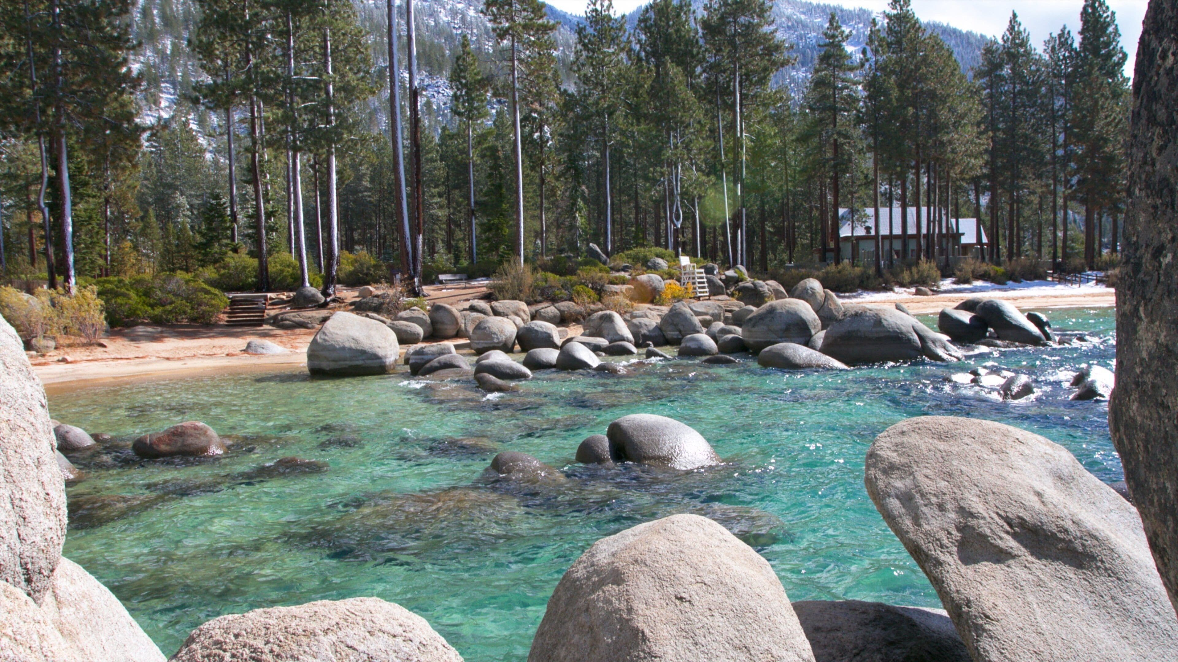 A clear river with large rocks, a sandy beach, and a forested mountain backdrop.